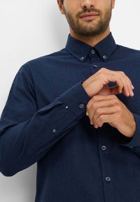 Camicia azzurra scuro con tessuto testurizzato, maniche lunghe e polsini con bottoni, dotata di un colletto tradizionale e bottoni frontali.