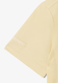 Manche courte d'une chemise en tissu perforé jaune pâle avec un petit logo blanc Lacoste sur le bord de la manche.