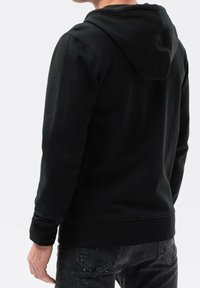 Personne portant un sweat à capuche noir et un jean foncé éclaboussé de peinture blanche, vue de dos sur un fond uni.