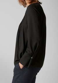 Blusa nera a maniche lunghe con pieghe sul davanti, tessuto morbido, dettagli con bottoni ai polsi, abbinata a pantaloni scuri con texture.
