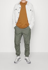 Muž oblečený v bílém fleecovém saku na knoflíky, pod kterým má hnědé tričko, olivově zelené teplákové kalhoty a bílo-šedé běžecké boty, s rukama v kapsách.