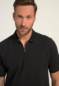 Camisa polo negra con cuello con cremallera, fabricada en un tejido suave. Presenta un logo sutil en relieve en el pecho y mangas cortas.