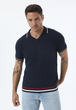Mand med tatoveringer iført marineblå kortærmet polo-shirt med røde og hvide striber samt lyseblå jeans, stående mod en hvid baggrund.