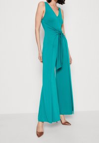 Monos de color turquesa con escote en V, corsé ajustado y detalle atado en la cintura, con piernas anchas y textura suave. Tacones marrones completan el look.