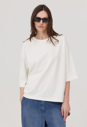 Kobieta nosząca czarne owalne okulary przeciwsłoneczne, luźną białą koszulę z krótkim rękawem i niebieskie dżinsy, stojąca na tle jednolitego jasnego tła.