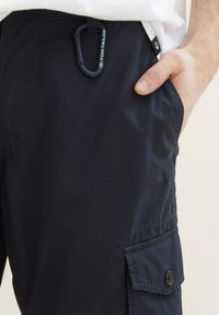 Dunkelblaue Cargo-Shorts aus strukturiertem Stoff, mit einer Seitentasche mit Druckknopfverschluss und einem Karabinerhaken, der an der Taille befestigt ist.
