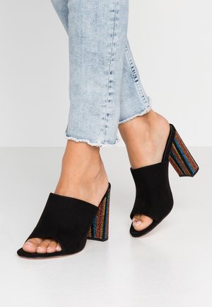 Femme portant un jean bleu clair et des mules noires à talons hauts ouverts avec des talons blocs pailletés à rayures multicolores.