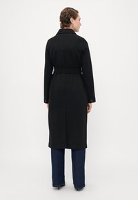 Persona con capelli corti vestita con un lungo cappotto nero con cintura, jeans blu scuri e scarpe con tacco nere, vista di spalle su uno sfondo neutro.