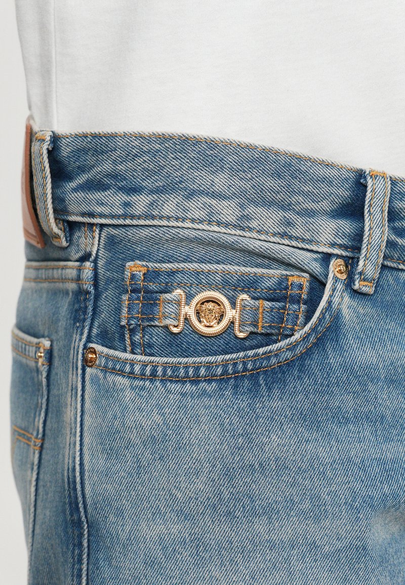Blå denimjeansficka med en gyllene dekorativ emblem med ett lejonhuvud på en liten myntficka, bärs med en vit skjorta.