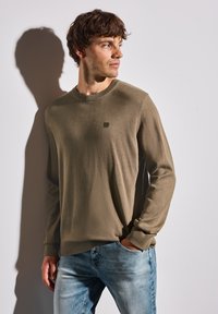 Ljus olivfärgad tröja med rund halsringning och ribbade muddar, med en liten logotyp på bröstet, bärs över blekta blå jeans.