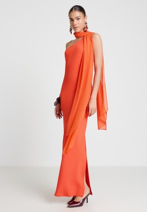 Femme en robe longue orange sans bretelles avec un détail d'écharpe fluide, talons hauts bordeaux et boucles d'oreilles remarquables, se tenant devant un fond uni.