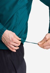 Mains ajustant le cordon de serrage à l'ourlet d'une veste verte portée sur un pantalon noir sur un fond uni.