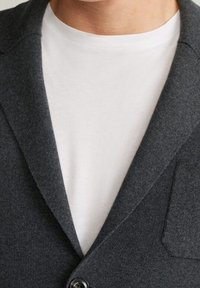 Persona che indossa una maglietta bianca a girocollo sotto un blazer grigio scuro con bottoni, con una tasca sul petto visibile.