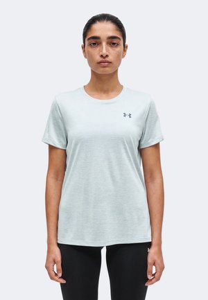 Sötét hajú nő világosszürke, rövid ujjú Under Armour pólóban és fekete leggingsben, egy egyszínű világosszürke háttér előtt áll.