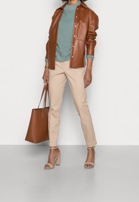 Kobieta w beżowych spodniach, beżowych sandałach na obcasie, niebieskiej swetrze w prążki, brązowej skórzanej kurtce, trzymająca dużą brązową torbę.