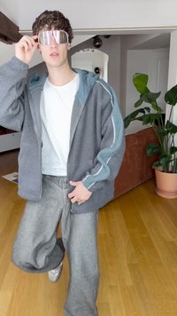 Grå fleecetröja med dragkedja och ljusblå detaljer, vit t-shirt, grå vida byxor och reflekterande rosa solglasögon. Rum med synliga växter.