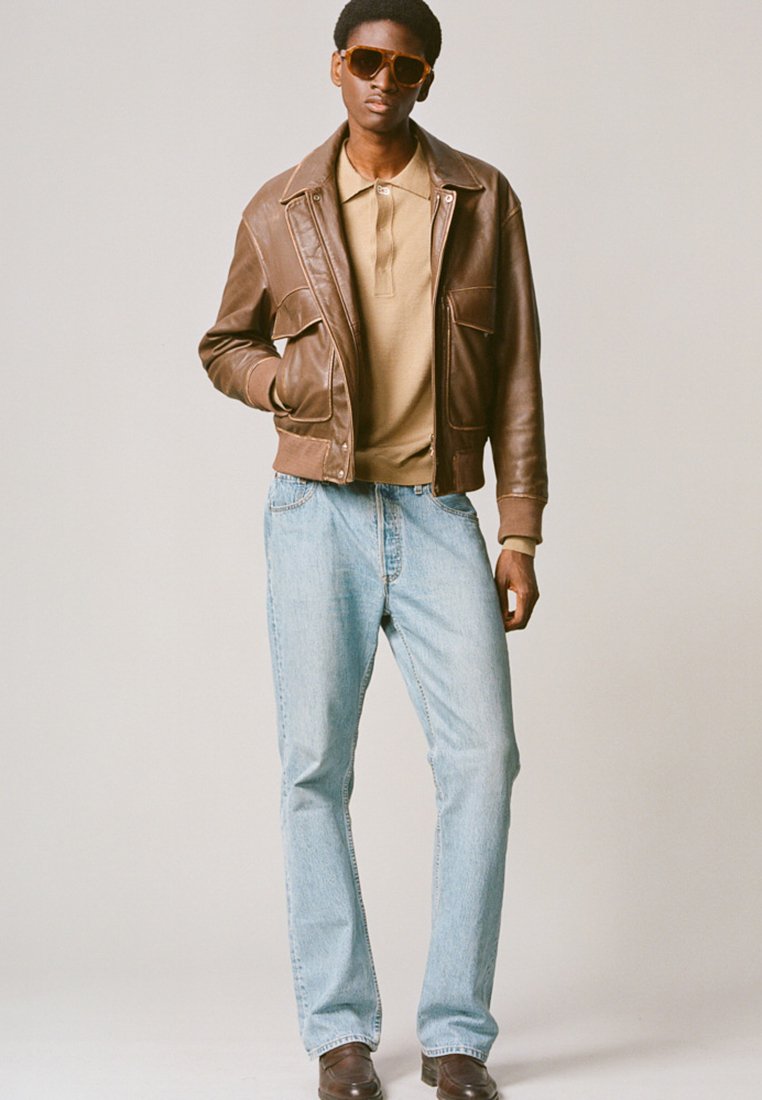 Giacca di pelle marrone sopra una polo beige, abbinata a jeans azzurri chiari e scarpe marroni. Grandi occhiali da sole marroni completano il look.