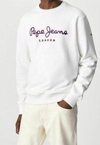 Man som bär en vit sweatshirt med "Pepe Jeans London"-logotyp i mörkröd färg, tillsammans med ljusbeiga byxor, med en hand i fickan.