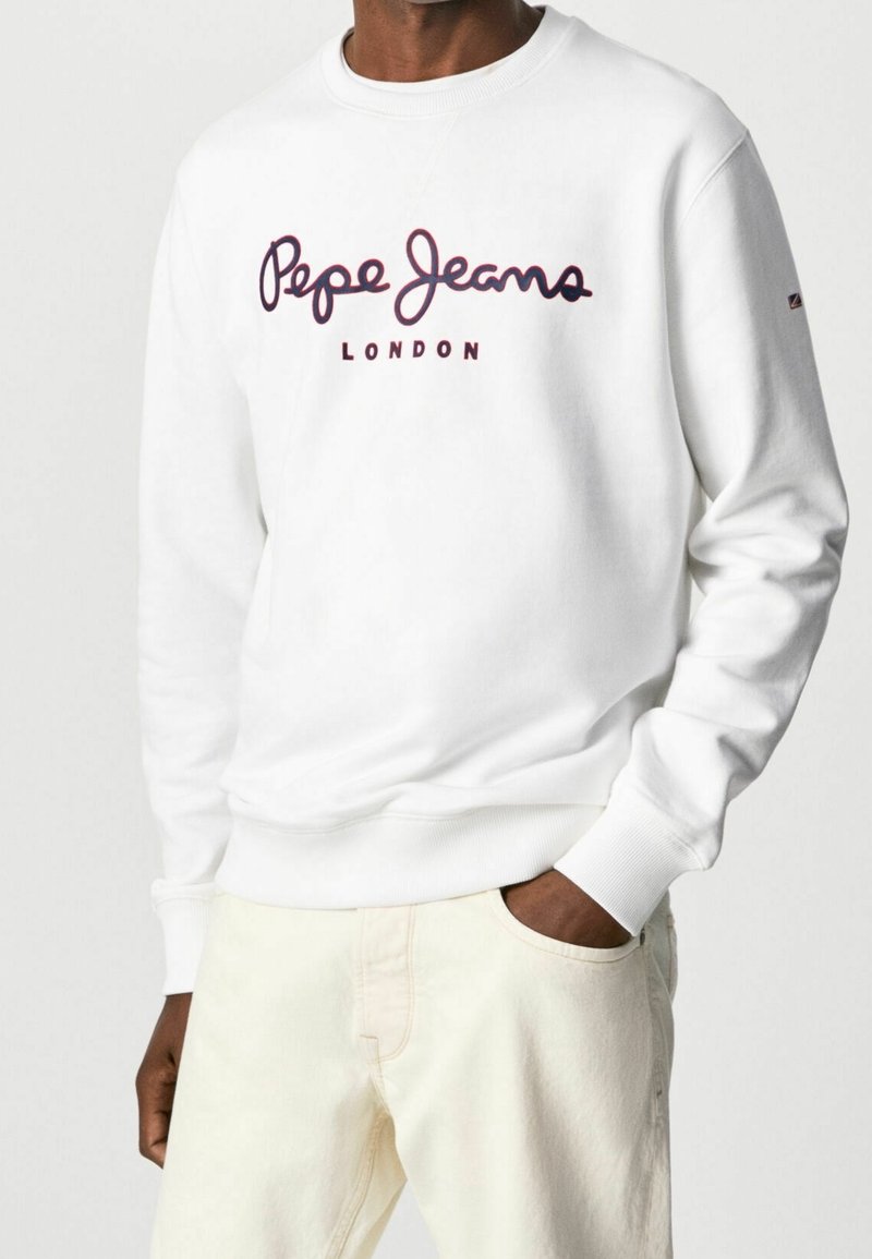 Man som bär en vit sweatshirt med "Pepe Jeans London"-logotyp i mörkröd färg, tillsammans med ljusbeiga byxor, med en hand i fickan.
