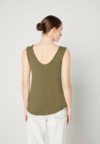Olive green sleeveless top with a wide back neckline, textured fabric, and curved hem, paired with light-colored loose-fitting pants.