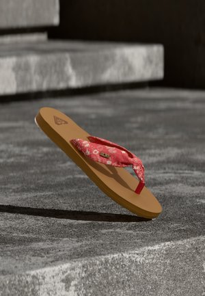 Chancla sencilla de color bronceado con una correa de tela roja que presenta patrones florales y el logo "Roxy", descansando sobre una superficie de concreto con escalones al fondo.