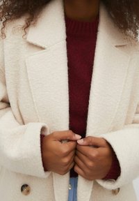 Person, der bærer en tekstureret lysecremefarvet frakke over en maroon sweater, som justerer frakkens front med begge hænder.