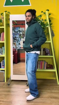 Green hooded sweatshirt, light jeans, and white sneakers. Stand-alone mirror and yellow bookshelf with books and plants in the background.