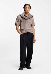 Mann trägt ein taupefarbenes, strukturiertes Kurzarmhemd mit passendem Pullover über den Schultern, schwarze, weit geschnittene Hose und schwarze Schuhe.