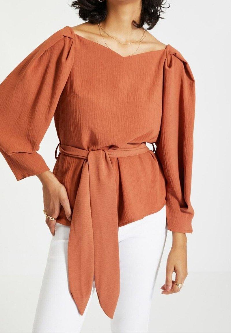 Femme portant un chemisier à manches longues couleur rouille avec des épaules bouffantes et une large ceinture à la taille, associé à un pantalon blanc.