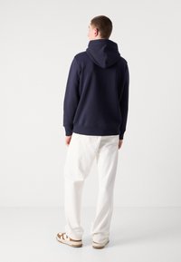 Sudadera azul marino con capucha ajustable, puños y dobladillo acanalados, combinada con pantalones blancos de corte holgado y calzado con detalles marrones.