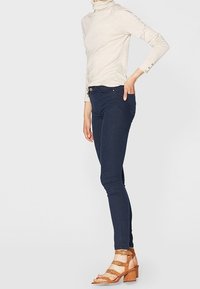 Jeans skinny azul oscuro con una textura suave, combinados con un suéter beige de cuello alto y sandalias de tacón strappy en color marrón claro. Sin patrones ni acentos notables.
