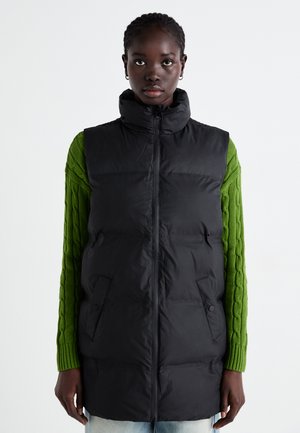 Gilet rembourré noir avec col montant et fermeture éclair à l'avant, doté de deux poches latérales. Porté par-dessus un pull en laine torsadée vert.