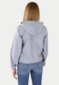 Chaqueta ligera de color gris claro con capucha, puños elásticos y corte recto, combinada con jeans de mezclilla azules.