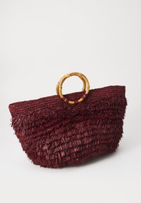 Piros szalmából készült tote táska texturált, rojtos dizájnnal és bambuszfüllel. A formája széles és sekély, alkalmas hétköznapi használatra.