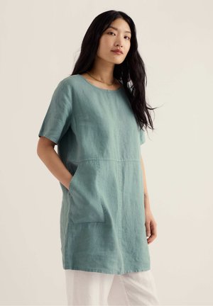 Kvinde med langt sort hår iført en løs teal tunika med lommer over hvide bukser, stående mod en ensartet lys baggrund.