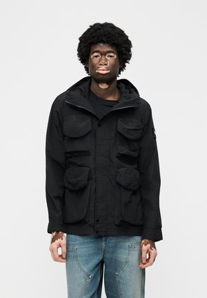 Fiatal vitiligós személy fekete, több zsebes dzsekit és világoskék farmert visel, egy sima fehér háttér előtt áll.