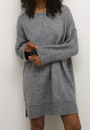 Kobieta w oversizowej szarej sukience swetrowej z długimi rękawami, stojąca na tle jasnego, jednolitego tła, poprawiająca mankiety splecionymi dłońmi.