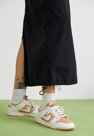 Ben iført hvide og tan Nike-sneakers med hvide sokker, sorte brede bukser med snøre i kanten, stående på et grønt gulv. Tatovering synlig på venstre ben.