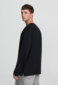 Czarna koszulka z długim rękawem o luźnym kroju, z okrągłym dekoltem i bocznymi rozcięciami. Model ma na sobie szare dżinsy; minimalistyczny design o gładkiej strukturze.