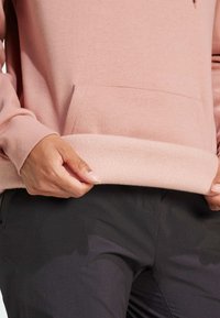 Felpa rosa con orlo interno in morbido pile, dotata di una tasca frontale. La persona indossa la felpa e tiene l'orlo, mostrando la texture contro i pantaloni scuri.