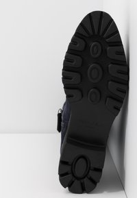 Semelle en caoutchouc noir d'une botte avec de larges crampons, présentant une finition texturée et un logo en relief sur le côté, attachée à une tige bleue marine.