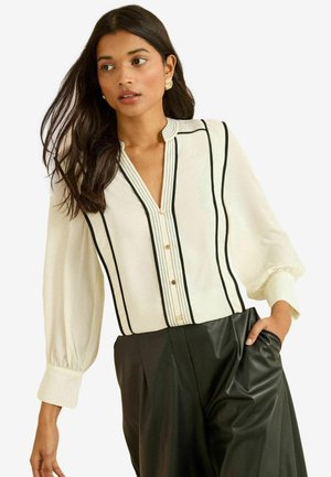 White blouse with black vertical stripes, button front, and a textured collar. Puffed sleeves and a relaxed fit paired with black pants.