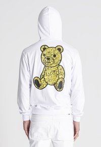 Biela mikina s kapucňou s veľkým žltým grafikom medveďa na chrbte, obkresleným čiernou farbou. Vyrobená z mäkkej tkaniny s prednou klokankou.