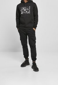 Felpa nera con testo grafico bianco, abbinata a pantaloni cargo neri con tasche laterali e polsini elastici. Sneakers nere completano l'outfit.
