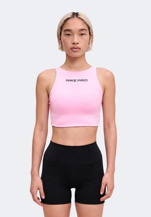 Jeune personne aux cheveux blonds courts portant un crop top rose Nike Pro et un short de sport noir taille haute, debout face à nous.