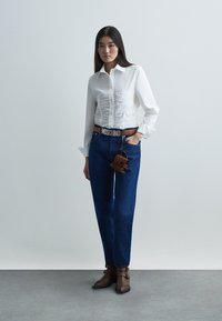 Λευκό εφαρμοστό πουκάμισο με ραβδώσεις στο μπροστινό μέρος, μπλε ψηλόμεσο τζιν, καφέ ζώνη με το λογότυπο «MOSCHINO», καφέ μποτάκια αστραγάλου και μικρή καφέ τσάντα χειρός.