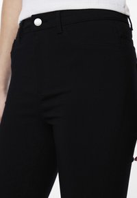 Primer plano de una persona que lleva unos vaqueros negros ajustados y una camiseta blanca, centrando la atención en la cintura y el área superior del muslo.