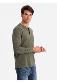Grünes Langarm-Henley-Shirt mit Rundhalsausschnitt, ausgestattet mit einer Knopfleiste vorne und seitlichen Schlitzen, getragen zu blauen Jeans.