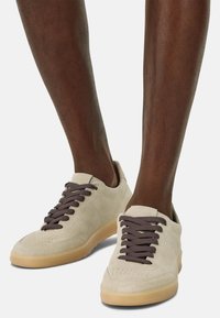 Beige suede sneakers with brown laces, rounded toe, and textured rubber sole, displaying a simple design and minimal branding.