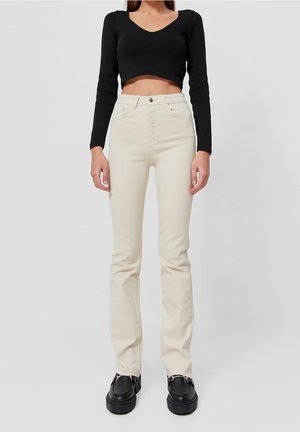 Femme portant un jean droit taille haute écru, un top noir court à manches longues et des mocassins à plateforme noirs, debout devant un fond uni.